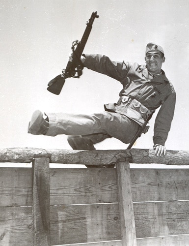 A man undergoing intense training at a boot camp, leaning over a fence with a rifle.