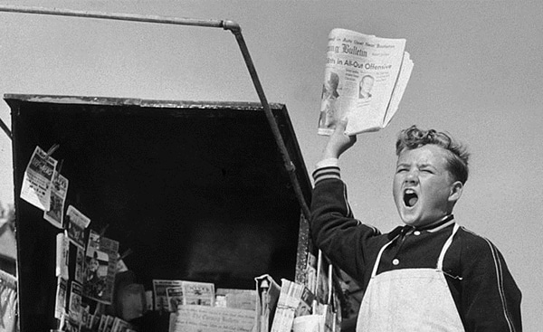A boy keeping up with the news, holding a newspaper in front of a truck.