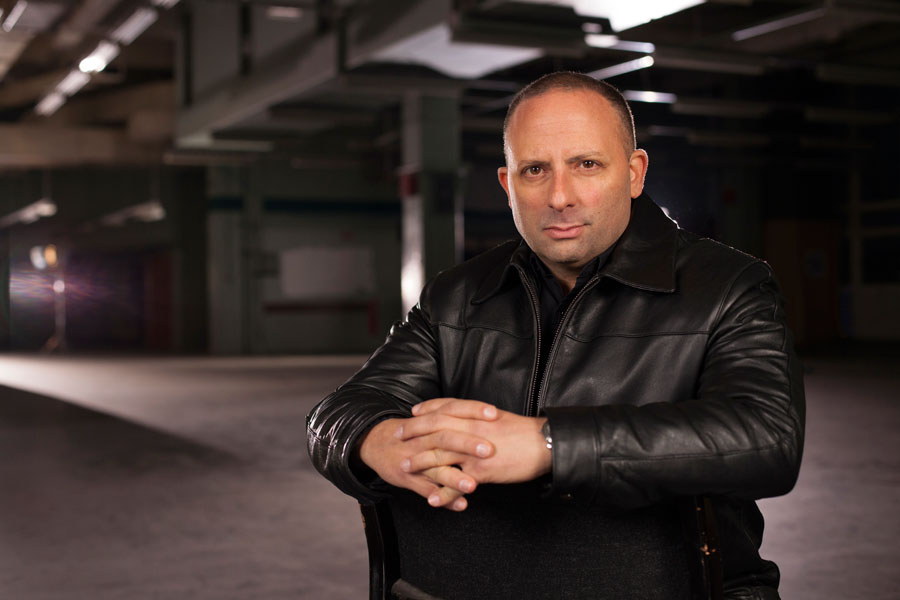 A man in a leather jacket sits on a chair in an empty building, possibly recording a new podcast episode.