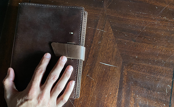 A person's hand holding a brown notebook on a wooden table, showcasing their love for journaling.