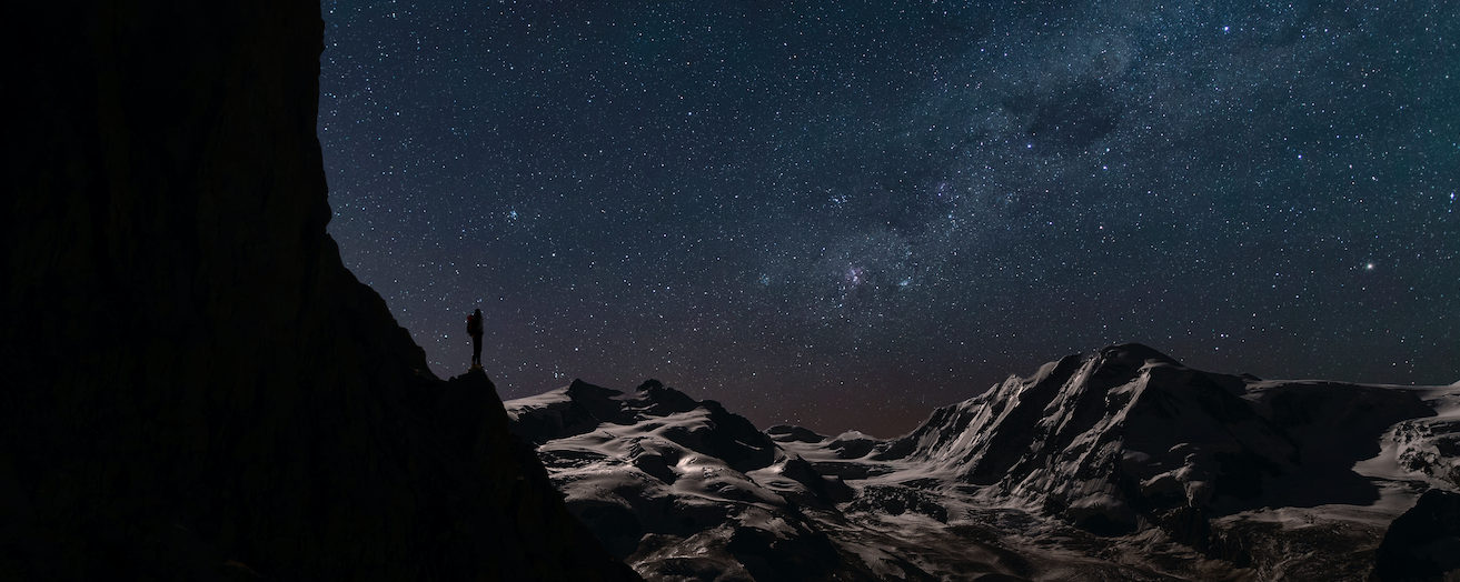 A person standing on top of a mountain with the milky way in the sky, contemplating existence.